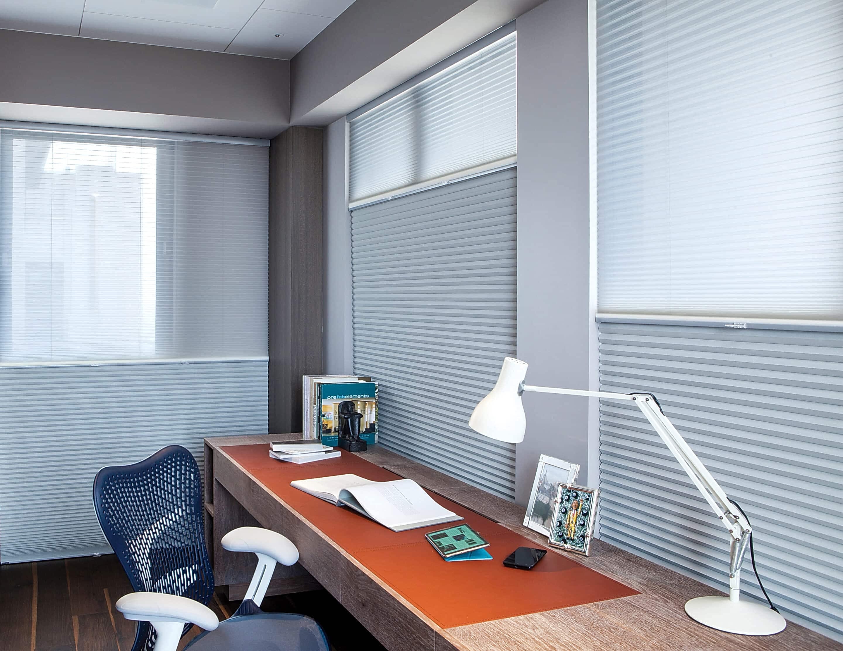 Modern home office with a desk, chair, and white lamp, featuring top-down bottom-up cellular shades on multiple windows.